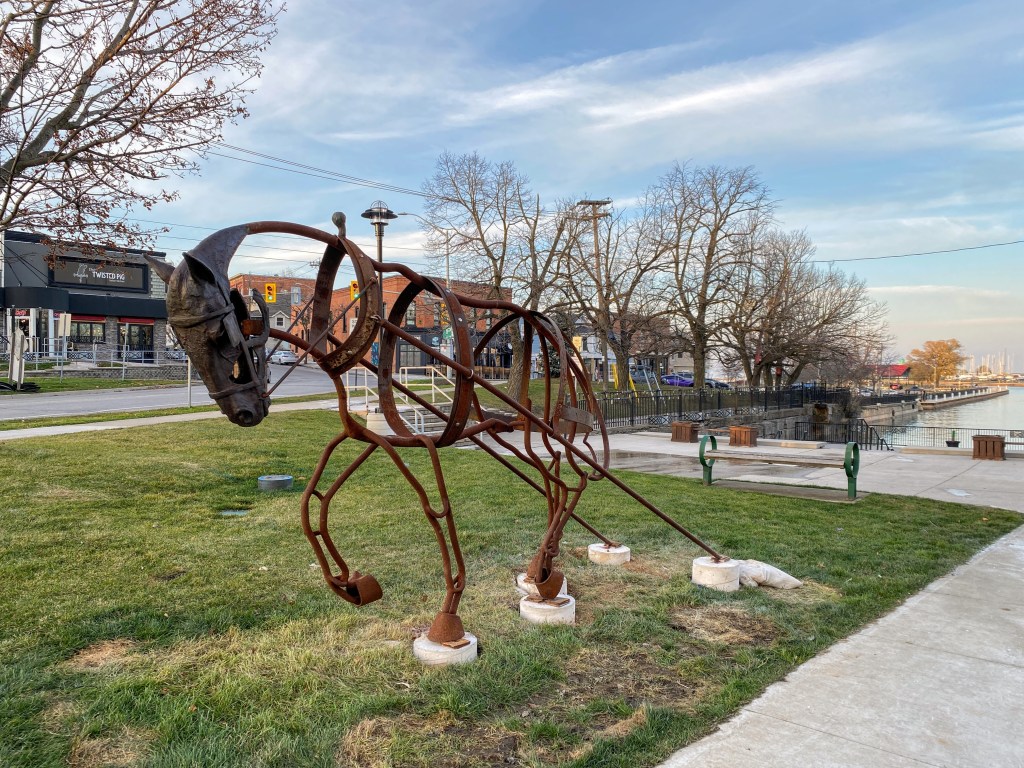A large metal sculpture of a workhorse on a patch of grass near the edge of Lake Ontario. There is a road and several shops in the background. 